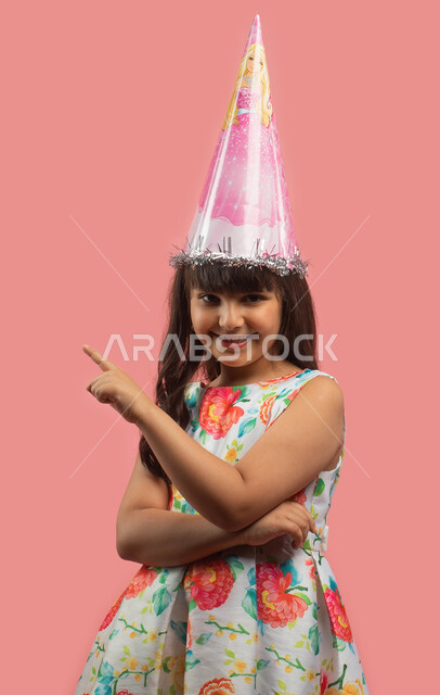 أعياد ومناسبات سعيدة، بورتريه لفتاة عربية خليجية سعودية ترتدي فستان مورد أنيق وقبعة الإحتفال بعيد الميلاد وتشير باصبع السبابة نحو شيء ما، حركة أصبع وإشارات، إيماءات وجه تدل على الفرح، خلفية وردية