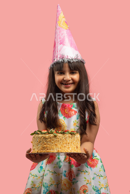قضاء أوقات ممتعة، اعياد ومناسبات سعيدة، بورتريه لفتاة عربية سعودية خليجية ترتدي قبعة الإحتفال بعيد الميلاد وتحمل قالب الكيك المزين بالفراولة، إيماءات وجه تدل على السعادة والفرح، خلفية وردية