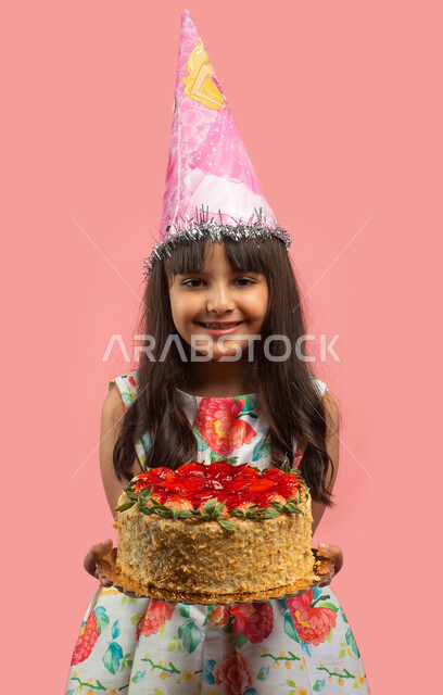 اعياد ومناسبات سعيدة، بورتريه لفتاة عربية سعودية خليجية ترتدي قبعة الإحتفال بعيد الميلاد وتحمل قالب الكيك المزين بالفراولة، قضاء أوقات ممتعة، إيماءات وجه تدل على الفرح، خلفية وردية
