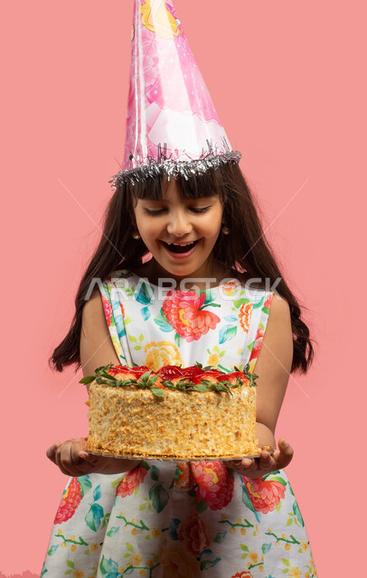 قضاء أوقات ممتعة، بورتريه لفتاة عربية سعودية خليجية ترتدي قبعة الإحتفال بعيد الميلاد وتحمل قالب الكيك المزين بالفراولة، إيماءات وجه تدل على الدهشة والاعجاب والفرح، اعياد ومناسبات سعيدة، خلفية وردية