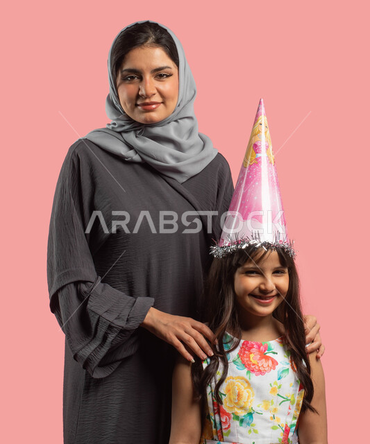 بورتريه لأم عربية خليجية سعودية تقف الى جانب ابنتها، إيماءات تدل على الحب والفرح، طفلة ترتدي ترتدي فستان مورد أنيق وقبعة الإحتفال بعيد الميلاد، أعياد ومناسبات سعيدة، خلفية وردية