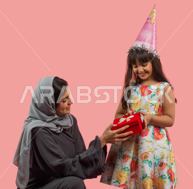 أمرأة مبتسمة تحتفل بعيد ميلاد طفلتها، بورتريه لأم عربية خليجية سعودية تقوم بإهداء إبنتها علبة هدايا حمراء، أعياد ومناسبات عائلية سعيدة، تقديم هدية يوم الميلاد، إيماءات وجه ويدين تدل على الفرح، زينة الحفلات والمناسبات، خلفية وردية