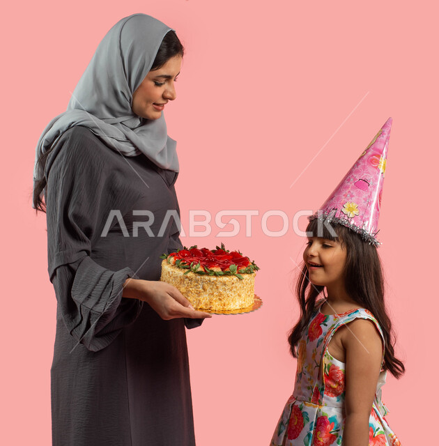 بورتريه لأم تقوم بتقديم قالب الكيك المزين بالفواكه لإبنتها، أمرأة عربية خليجية سعودية مبتسمة تحتفل بعيد ميلاد طفلتها، أعياد ومناسبات عائلية سعيدة، إيماءات وجه تدل على الإعجاب والفرح، تقديم هدية يوم الميلاد، خلفية وردية