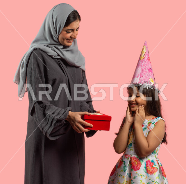 أم تفاجئ إبنتها بعلبة هدايا حمراء، بورتريه لأمرأة عربية خليجية سعودية مبتسمة تحتفل بعيد ميلاد طفلتها، أعياد ومناسبات عائلية سعيدة، تقديم هدية يوم الميلاد، إيماءات وجه ويدين تدل على الدهشة والمفاجئة والفرح، خلفية وردية