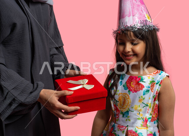 Giving a birthday gift, party and event decorations, a portrait of a Saudi Arabian Gulf mother giving her daughter a red gift box, a smiling woman celebrating her daughter’s birthday, happy family holidays and occasions, face and hand gestures indicating 
