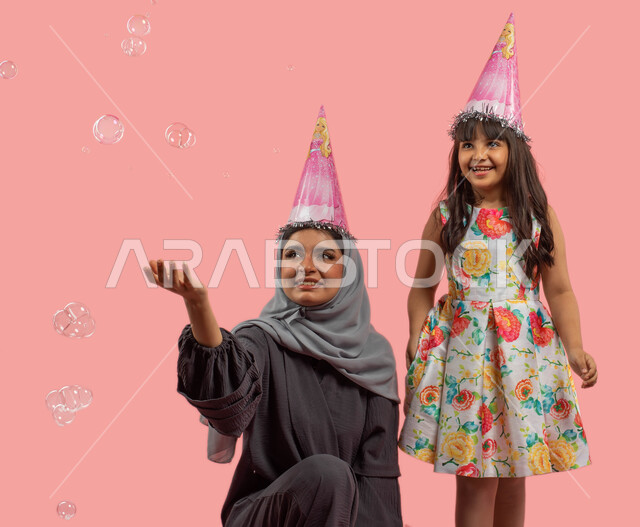 اللعب بالفقاعات المائية المتطايرة والمتناثرة، أعياد ومناسبات عائلية سعيدة، بورتريه لأم عربية خليجية سعودية تردي قبعة الاحتفال بعيد الميلاد مع ابنتها، قضاء الأوقات في الأنشطة الترفيهية الممتعة، إيماءات وجه تدل على الإستمتاع والفرح، خلفية وردية