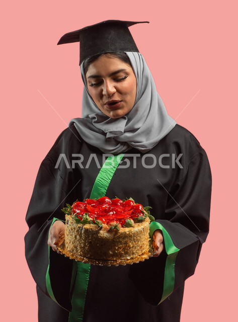 النجاح والتفوق والتخرج من الجامعة، خريجة تحمل قالب الكيك المزين بالفواكه، بورتريه لطالبة جامعية عربية سعودية خليجية ترتدي العباءة وقبعة التخرج، أجواء إحتفالية سعيدة، التعليم في المملكة العربية السعودية، إيماءات وجه تدل على السعادة، خلفية وردية