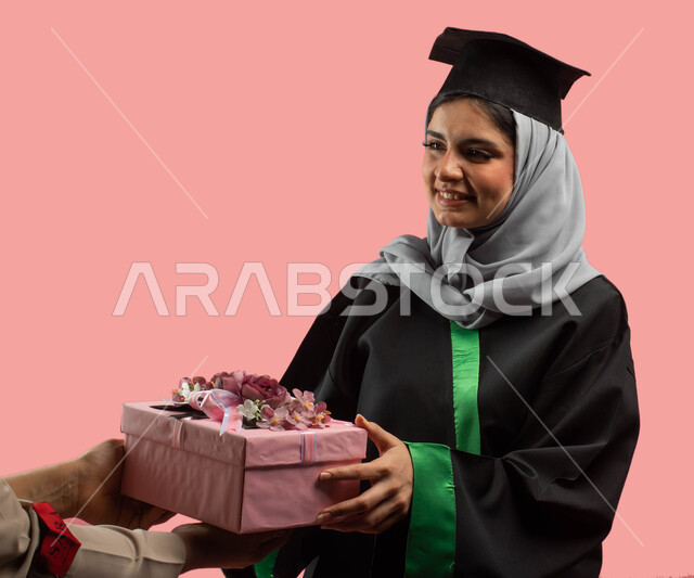 Education in the Kingdom of Saudi Arabia, a portrait of a Saudi Arabian Gulf university graduate wearing an abaya and a graduation hat and holding a closed pink gift box in her hand, success, excellence and graduation from the university, facial gestures 