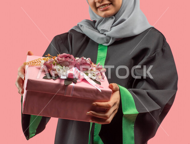 النجاح والتفوق والتخرج من الجامعة، بورتريه مقرب لخريجة جامعية عربية سعودية خليجية ترتدي العباءة وقبعة التخرج وتحمل بيدها علبة هدايا زهرية مغلفة، أجواء إحتفالية سعيدة، إيماءات وجه تدل على السعادة، التعليم في المملكة العربية السعودية، خلفية وردية