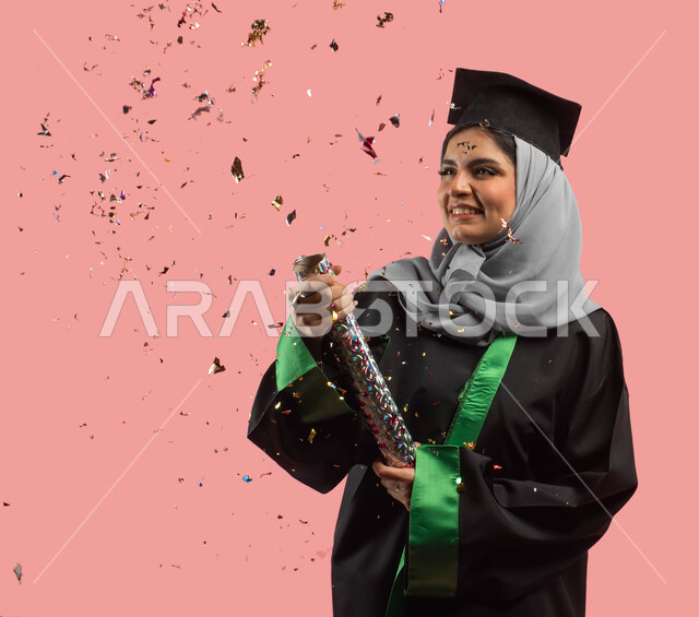 استخدام مفرقعات الاحتفالات الورقية، بورتريه لطالبة جامعية عربية سعودية خليجية ترتدي العباءة وقبعة التخرج، التعليم في المملكة العربية السعودية، خريجة تحتفل بالنجاح والتفوق والتخرج من الجامعة، ايماءات وجه تدل على السعادة، خلفية وردية