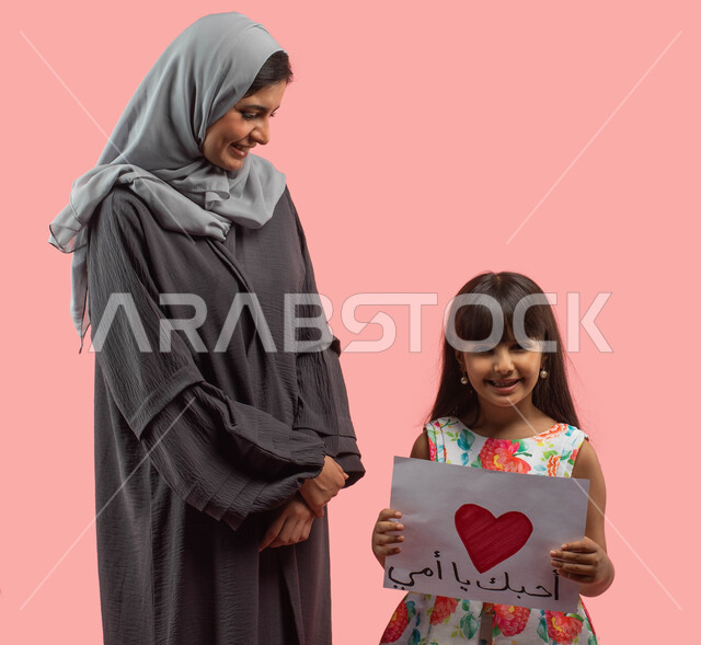 مفهوم حب الأم والإحسان إليها، بورتريه لطفلة عربية خليجية سعودية تعطي والدتها بطاقة معايدة ذات قلب احمر وكلمة أحبك يا أمي في عيد الام، اعياد ومناسبات سعيدة، الاحتفال بعيد الميلاد، تقديم هدية يوم الام، ايماءات تدل على الإعجاب والفرح، خلفية وردية
