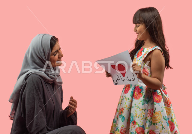 Happy holidays and occasions, a portrait of a Saudi Arabian Gulf girl giving her mother a greeting card with a red heart and the word “I love you, Mom” on Mother’s Day, celebrating Christmas, presenting a Mother’s Day gift, gestures indicating admiration 