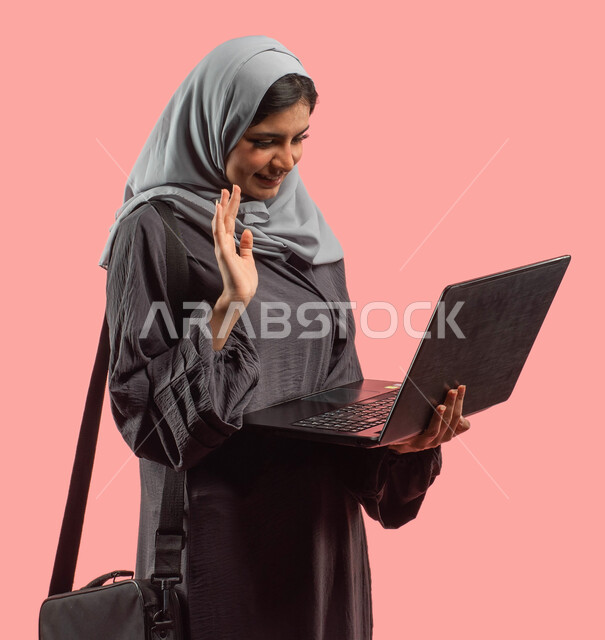 إجراء مكالمات فيديو عبر الإنترنت، بورتريه لإمرأة عربية خليجية تحمل بيدها جهاز الحاسوب المحمول وتلوح بيدها، إيماءات وجه ويدين تدل على التحية والترحيب، إنجاز مهام العمل عن بعد، إستخدام وسائل التواصل الإجتماعي، خلفية وردية