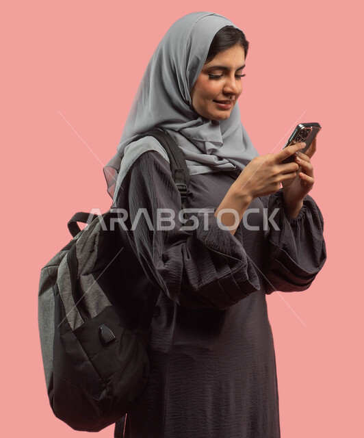 Searching and reading through a mobile phone, a portrait of a Saudi Gulf Arab university student carrying a backpack and using a mobile phone, browsing applications and social media, preparing for the new academic year, a pink background
