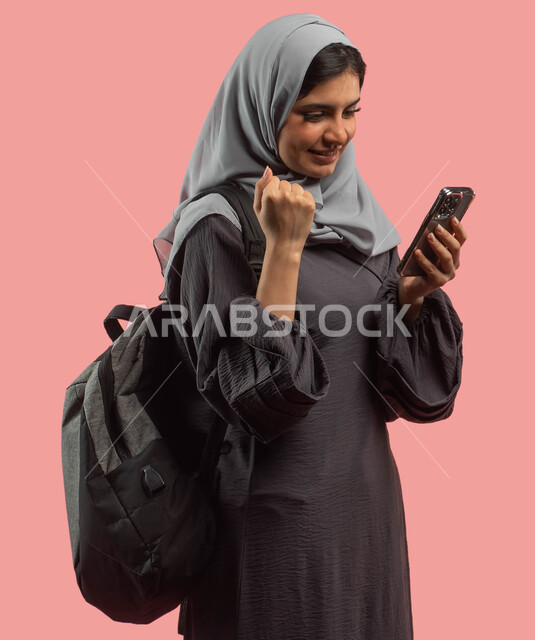 متابعة الدرجات من خلال الجوال، بورتريه لطالبة جامعية عربية خليجية سعودية تحمل حقيبة الظهر وتستخدم الهاتف المحمول، البحث والمطالعة وتصفح التطبيقات الالكترونية، الاستعداد للعام الدراسي الجامعي الجديد، ايماءات الوجه واليد تدل على النجاح والامتياز، خلفية وردي