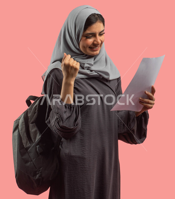 النجاح في الإختبار وإجتياز المرحلة الدراسية، بورتريه لطالبة جامعية عربية خليجية سعودية تحمل حقيبة ظهر وتمسك ورقة الإختبارات، إيماءات وجه ويدين تدل على النجاح والإمتياز، التعليم في المملكة العربية السعودية، خلفية وردية