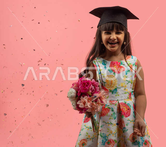 التخرج من المدرسة، مناسبات وأجواء إحتفالية سعيدة، بورتريه لفتاة عربية خليجية سعودية ترتدي فستان مورد وقبعة التخرج وتحمل بيدها باقة أزهار، إيماءات وجه تدل على الحماس والسعادة بالنجاح والتعلم، زينة ومفرقعات الحفلات، خلفية وردية