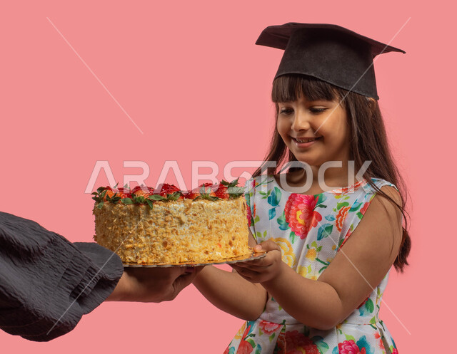 التخرج من المدرسة، قالب الكيك المزين بالفواكه، بورتريه لفتاة عربية خليجية سعودية ترتدي فستان مورد وقبعة التخرج، مناسبات وأجواء إحتفالية سعيدة، إيماءات وجه تدل على السعادة بالنجاح، خلفية وردية