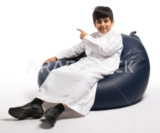 إيماءات وتعابير وجه تدل على الإعجاب بشيء جميل , بورتريه لطفل عربي سعودي خليجي يرتدي الزي السعودي يجلس على كرسي مريح و يرفع يده للأعلى, الإشارة الى شيء ما , خلفية بيضاء