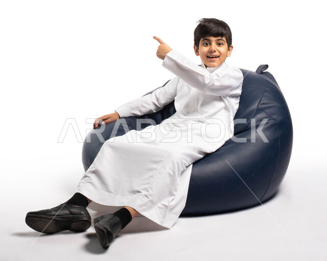 الإشارة الى شيء ما , إيماءات وتعابير وجه تدل على رؤية شيء جميل و الإعجاب به , بورتريه لولد عربي سعودي خليجي يرتدي الثوب التقليدي يجلس على أريكة مريحة و يرفع يده للأعلى, خلفية بيضاء