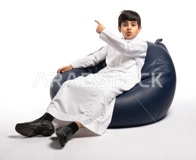 إيماءات وتعابير وجه تدل على التفاجؤ و الدهشة , بورتريه لصبي عربي سعودي خليجي يرتدي الثوب السعودي يجلس على كرسي مريح و يرفع يده للأعلى, الإشارة الى شيء ما , خلفية بيضاء