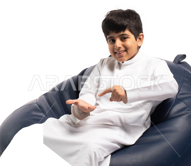 إيماءات وتعابير وجه تدل على الإعجاب بالمنتج, بورتريه لطفل عربي سعودي خليجي يرتدي الزي السعودي يجلس على كرسي مريح يحمل بيده شيء ما ويشير بإصبع السبابة ليده الأخرى, خلفية بيضاء