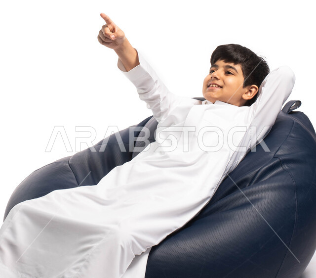 إيماءات وتعابير وجه تدل على رؤية شيء جميل , بورتريه لطفل عربي سعودي خليجي يرتدي الزي السعودي يجلس على كرسي مريح و يرفع يده للأعلى, الإشارة الى شيء ما , خلفية بيضاء