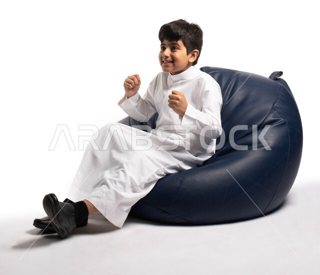 Spending fun times by watching the matches, gestures of the face and hands indicating enjoyment and enthusiasm in watching the events of the matches, a portrait of a Saudi Arabian Gulf child wearing the Saudi uniform sitting on a comfortable chair and wat