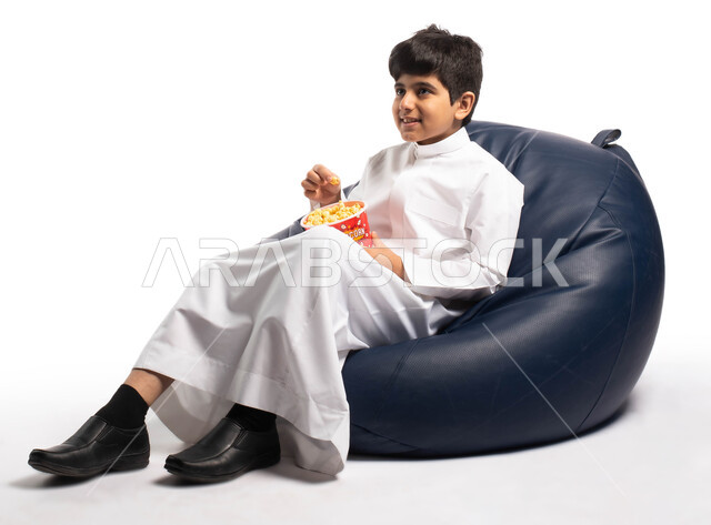  تمضية أوقات رائعة بمتابعة الأفلام، ايماءات وجه تدل على الاستمتاع برؤية فلم سينيمائي, بورتريه لطفل عربي خليجي سعودي يجلس على كرسي مريح يحمل بيده الفشار ، التسليه والترفيه بتناول دلو الفشار، خلفية بيضاء