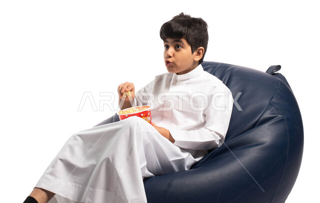 ايماءات وجه تعبر عن الاندهاش، قضاء وقت ممتع برؤية فيلم سينمائي، الاستمتاع بتناول الفشار, بورتريه لولد عربي خليجي سعودي يجلس على كرسي مريح يقوم بتناول الفشار ومشاهدة الأفلام، خلفية بيضاء