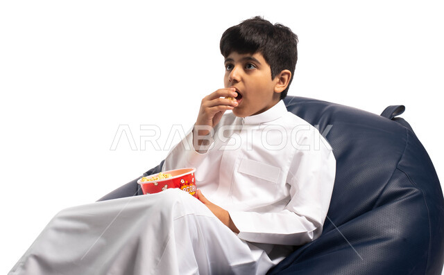 قضاء وقت ممتع برؤية فيلم سينمائي، الاستمتاع بتناول الفشار, بورتريه لطفل عربي خليجي سعودي يجلس على كرسي مريح يقوم بتناول الفشار ومشاهدة الأفلام، ايماءات وجه تعبر عن الاندهاش، خلفية بيضاء
