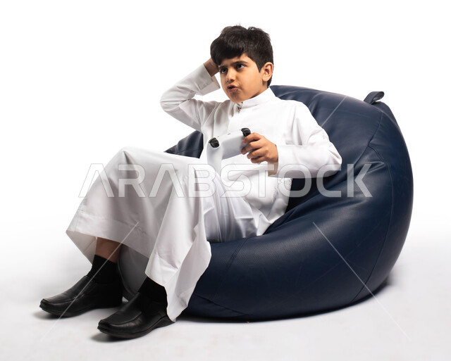 ايماءات وتعابير وجه و يدين تدل على صعوبة اللعب والفوز, بورتريه لطفل عربي خليجي سعودي يرتدي الزي السعودي يجلس على كرسي مريح ويمسك بيده يد التحكم و يلعب بألعاب الفيديو ، المتعة و التسلية بألعاب الفيديو و البلايستيشن الترفيهية، خلفية بيضاء