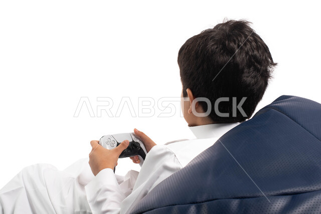 المتعة و التسلية بألعاب الفيديو و البلايستيشن الترفيهية , بورتريه من الخلف لطفل عربي خليجي سعودي يرتدي الزي السعودي ويجلس على كرسي مريح، يمسك بيده عصا التحكم و يلعب بألعاب الفيديو ، خلفية بيضاء