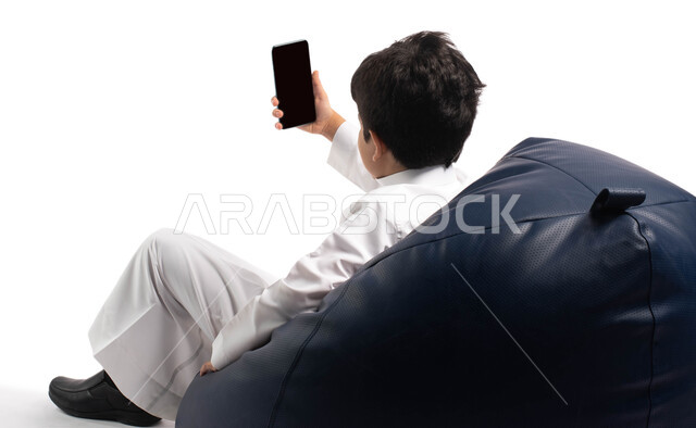 اجراء مكالمة فيديو والتواصل عن بعد , بورتريه من الخلف لطفل عربي خليجي سعودي يجلس على كرسي مريح و يحمل بيده هاتف محمول , إيماءات و تعابير وجه تدل على السعادة ، أخذ صورة سيلفي تذكارية ، خلفية بيضاء