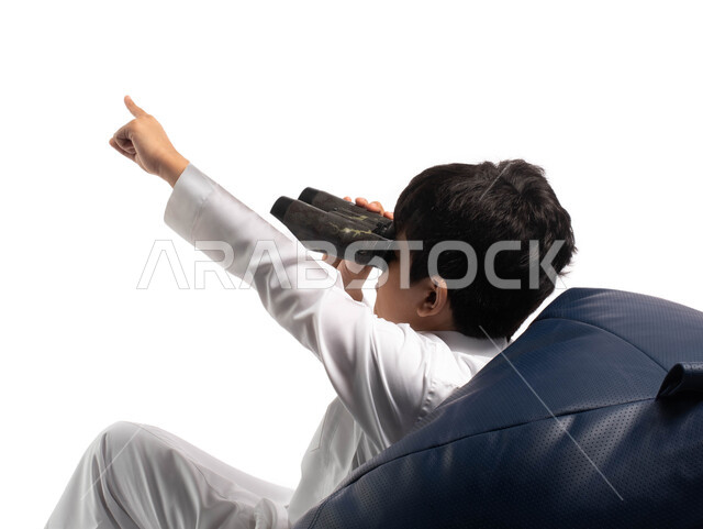 A future view of children, a portrait from behind of a Saudi Arabian Gulf boy wearing the traditional Saudi dress, sitting on a comfortable chair, holding binoculars in his hand, and pointing upwards, love of exploration and adventure, white background