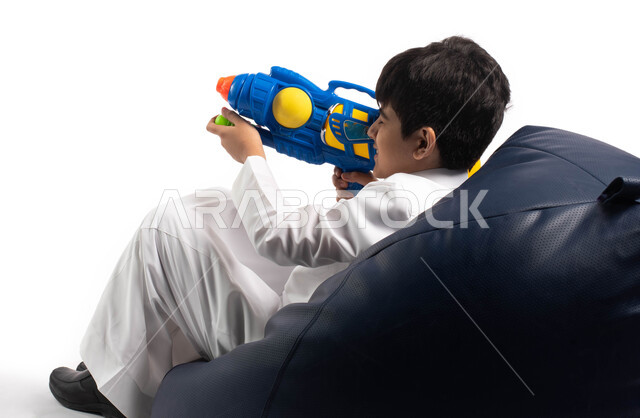  الاستمتاع والتسلية بأوقات الفراغ, مفهوم الترفيه عن النفس , بورتريه من الخلف لطفل عربي سعودي خليجي يجلس على كرسي مريح مستمتع باللعب بمسدس مائي بلاستيكي، العاب مائية في فصل الصيف ، أنشطة ترفيهية صيفية، خلفية بيضاء