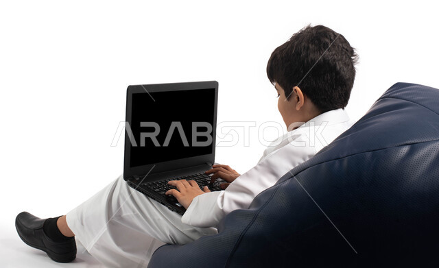 التواصل مع الأصدقاء عن بعد, محادثات ودردشة عبر الانترتت, بورتريه من الخلف لصبي عربي سعودي خليجي يرتدي الزي السعودي ويجلس على أريكة مريحة يقوم بإستخدام جهاز الحاسوب المحمول، مفهوم التعلم اونلاين، قضاء أوقات ممتعة على اللابتوب