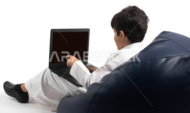  محادثات ودردشة عبر الانترتت, بورتريه من الخلف لطفل عربي سعودي خليجي يرتدي الزي السعودي ويجلس على كرسي مريح يقوم بإستخدام جهاز الحاسوب المحمول، التواصل مع الأصدقاء عن بعد، مفهوم التعلم اونلاين، قضاء أوقات ممتعة على اللابتوب