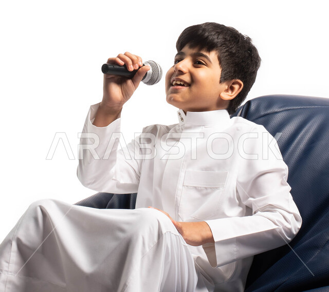 مفهوم تنمية المواهب الفنية, موهبة إلقاء الخطاب، بورتريه لصبي عربي سعودي خليجي يجلس على كرسي مريح و يحمل بيده الميكروفون، ايماءات وتعابير وجه تدل على الإستمتاع بالغناء، تقديم فقرات ترفيهية، تسجيل مقطع صوتي، تقديم العروض المسرحية، خلفية بيضاء