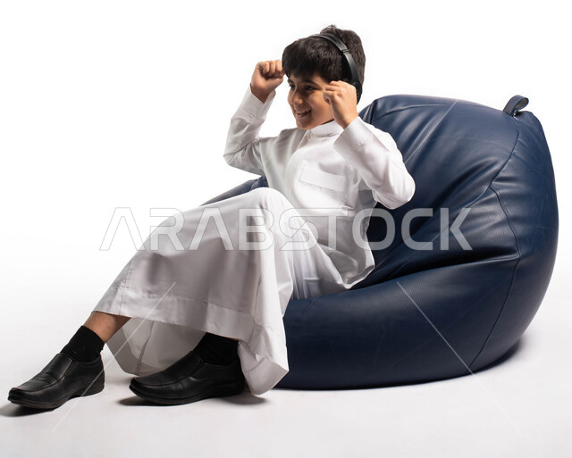 التسلية و الترفيه باستخدام تقنية الهيدفون, بورتريه صبي عربي سعودي خليجي يرتدي اللباس السعودي يجلس على كرسي مريح ويضع سماعات الرأس اللاسلكية ، ايماءات وجه ويدين تتعبر عن السعادة و الاستماع بحماس للأغاني ، خلفية بيضاء 