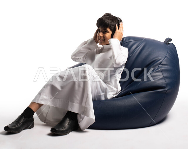 ايماءات وجه ويدين تدل على التركيز بسماع الأغاني ، المتعة و الترفيه باستخدام تقنية الهيدفون, بورتريه ولد عربي سعودي خليجي يرتدي الزي السعودي يجلس على كرسي مريح ويضع سماعات الرأس ، خلفية بيضاء 