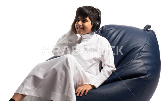 Leisure and entertainment using the headphone technology, a portrait of a Saudi Arabian Gulf child wearing the Saudi dress sitting on a comfortable chair and wearing headphones, facial gestures indicating happiness and enjoying listening to songs, a white