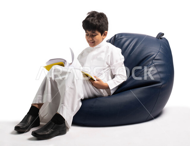 ثقافة التعليم الذاتي, الاستمتاع بقراءة الكتب, قضاء الوقت بالقراءة والتعلم والتثقف، ايماءات وتعابير وجه تدل على الاستمتاع بالمطالعة، بورتريه لطفل عربي سعودي خليجي يرتدي الثوب السعودي يجلس على كرسي مريح ويحمل بيديه كتاب, خلفية بيضاء