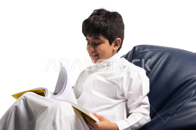 الاستمتاع بقراءة الكتب, قضاء الوقت بالقراءة والتعلم والتثقف، ايماءات وتعابير وجه تدل على الاستمتاع بالمطالعة، بورتريه لولد عربي سعودي خليجي يرتدي الزي السعودي يجلس على أريكة مريحة ويحمل بيديه كتاب, ثقافة التعليم الذاتي, خلفية بيضاء