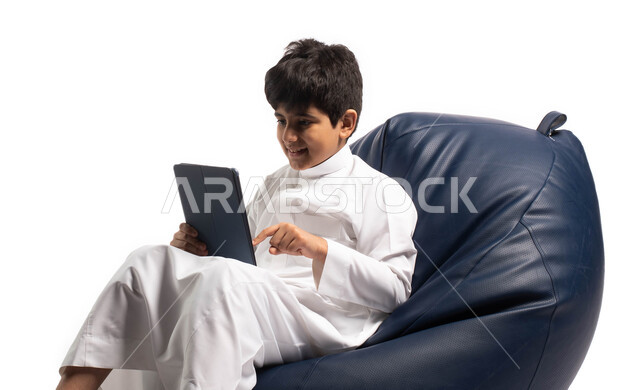 مفهوم التعليم عن بعد, اللعب على الجهاز المحمول, بورتريه لطفل عربي سعودي خليجي يرتدي اللباس السعودي يجلس على أريكه مريحه ويقوم باستخدام التابلت, ايماءات وتعابير وجه تدل على الاستمتاع والسعادة باستخدام التابلت, تسليه وترفيه على الأجهزة المحمولة, خلفية بيضاء