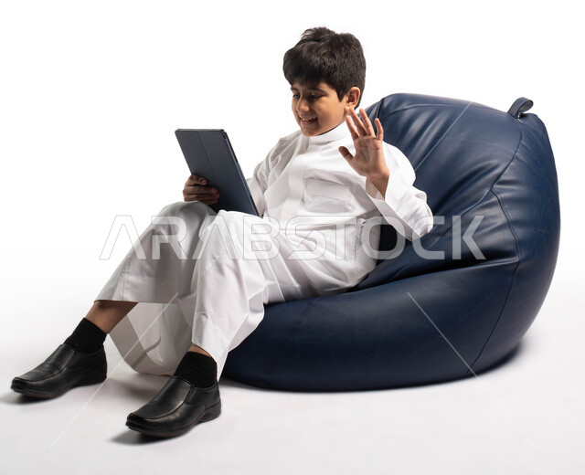 ايماءات وتعابير يد تدل على القاء التحية, بورتريه لولد عربي سعودي خليجي يرتدي الثوب السعودي يجلس على كرسي مريح ويقوم بالتواصل عبر التابلت,مفهوم التعليم عن بعد, تسليه وترفيه على الأجهزة المحمولة, قضاء وقت ممتع على الانترنت, خلفية بيضاء