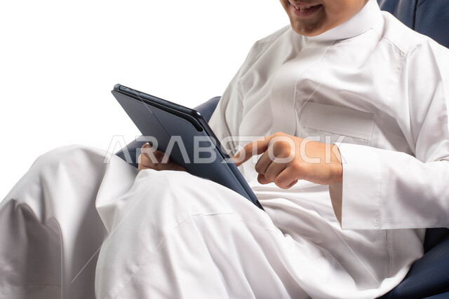 اللعب على الجهاز المحمول, بورتريه لطفل عربي سعودي خليجي يرتدي الزي السعودي يجلس على أريكه مريحه ويقوم باستخدام التابلت, انجاز المهام عبر التابلت, مفهوم التعليم عن بعد, تسليه وترفيه على الأجهزة المحمولة, قضاء وقت ممتع على الانترنت, خلفية بيضاء