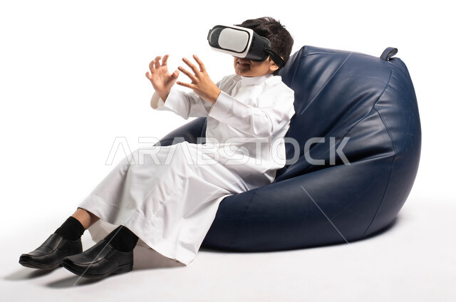 أنشطة ترفيهية للأطفال, بورتريه لطفل عربي خليجي سعودي يجلس على كرسي مريح يرتدي نظارة الواقع الإفتراضي، إيماءات و تعابير يدين تدل على السعادة والاستمتاع، المتعة و التسلية بمشاهدة الأفلام و ألعاب العالم الافتراضي ، تقنيات و ألعاب افتراضية، خلفية بيضاء