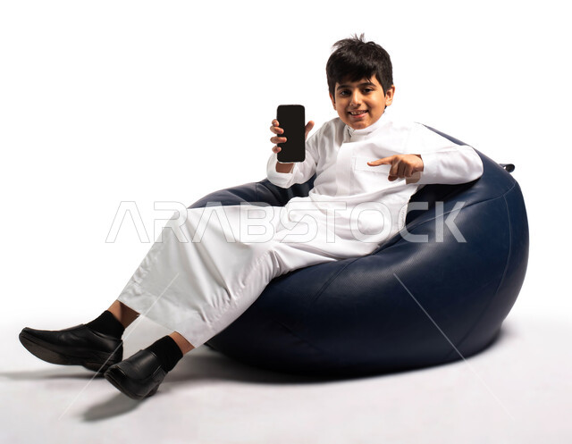 عرض خدمات وبرامج الهاتف المحمول, بورتريه لطفل عربي خليجي سعودي يرتدي الزي السعودي يجلس على كرسي مريح ويشير بيده الى الهاتف المحمول بشاشة سوداء فارغة، إستخدام أجهزة تقنية حديثة، مفهوم الاعلانات و التسوق الالكتروني, خلفية بيضاء