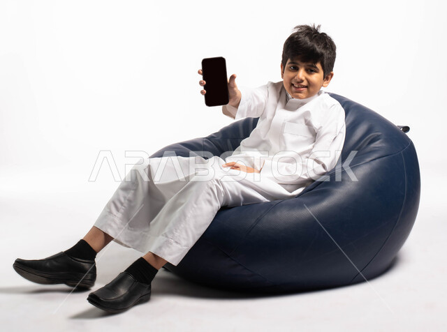 عرض خدمات وتطبيقات الهاتف المحمول, بورتريه لولد عربي خليجي سعودي يرتدي الزي السعودي يجلس على كرسي مريح و يمسك بيده الهاتف المحمول بشاشة سوداء فارغة، إستخدام أجهزة تقنية حديثة، مفهوم الاعلانات و التسوق الالكتروني, خلفية بيضاء